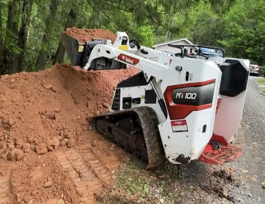 Mini track loader with a bucket of red clay working on a clay pile beside a road