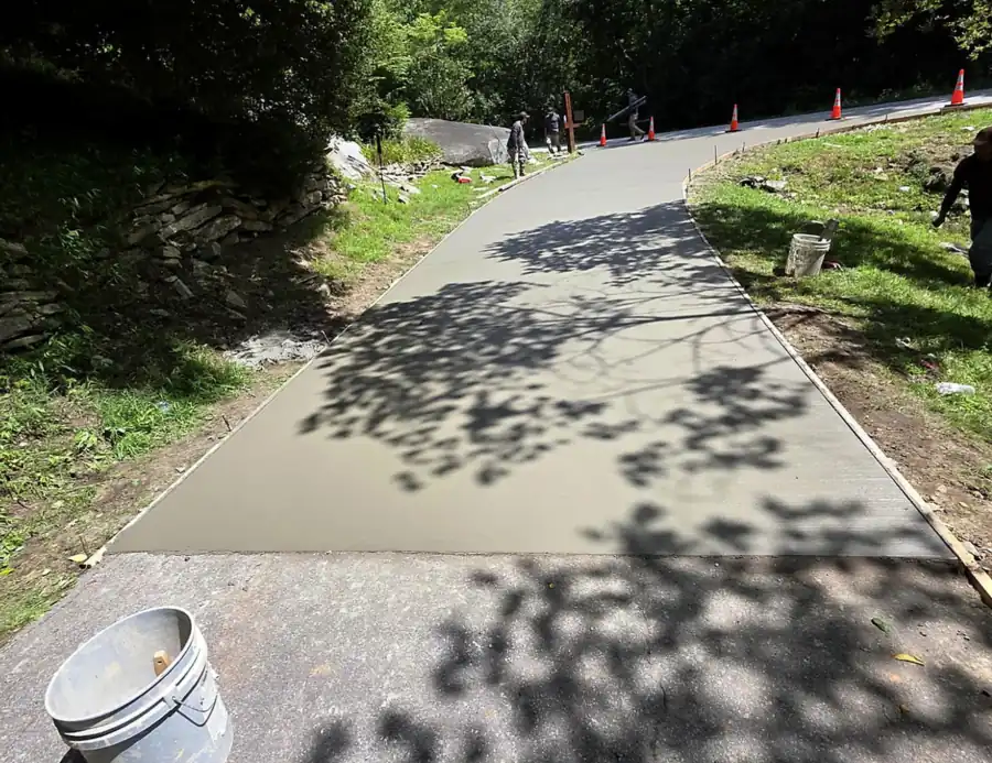 Fresh broom-finished concrete driveway curving uphill through trees, with cones and workers on it