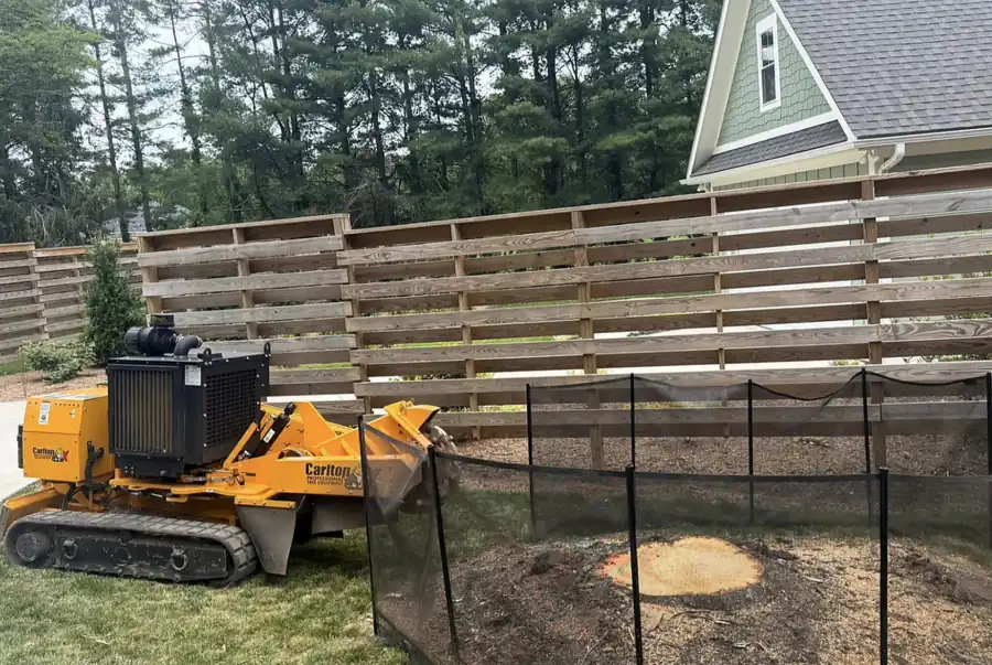 Tracked stump grinder and mesh debris screen set up at a low cut stump in front of a horizontal board fence