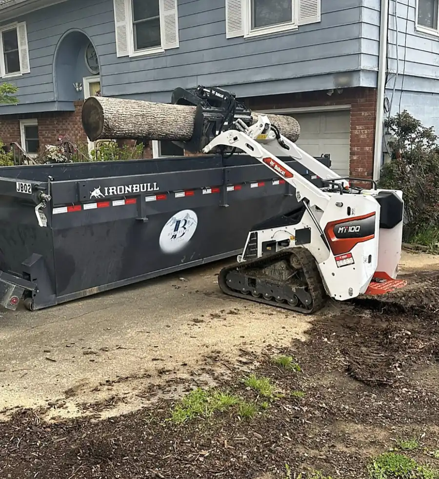 Mini track loader with a grapple lifting a large log into a roll-off bin beside a house