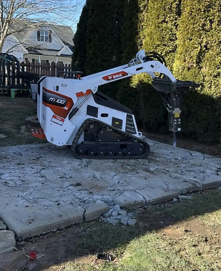 Mini track loader fitted with a hydraulic breaker breaking up a concrete slab beside a hedge