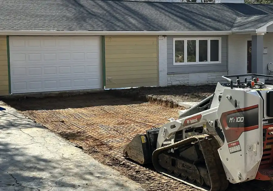 Mini track loader with a bucket beside a freshly graded pad in front of a garage
