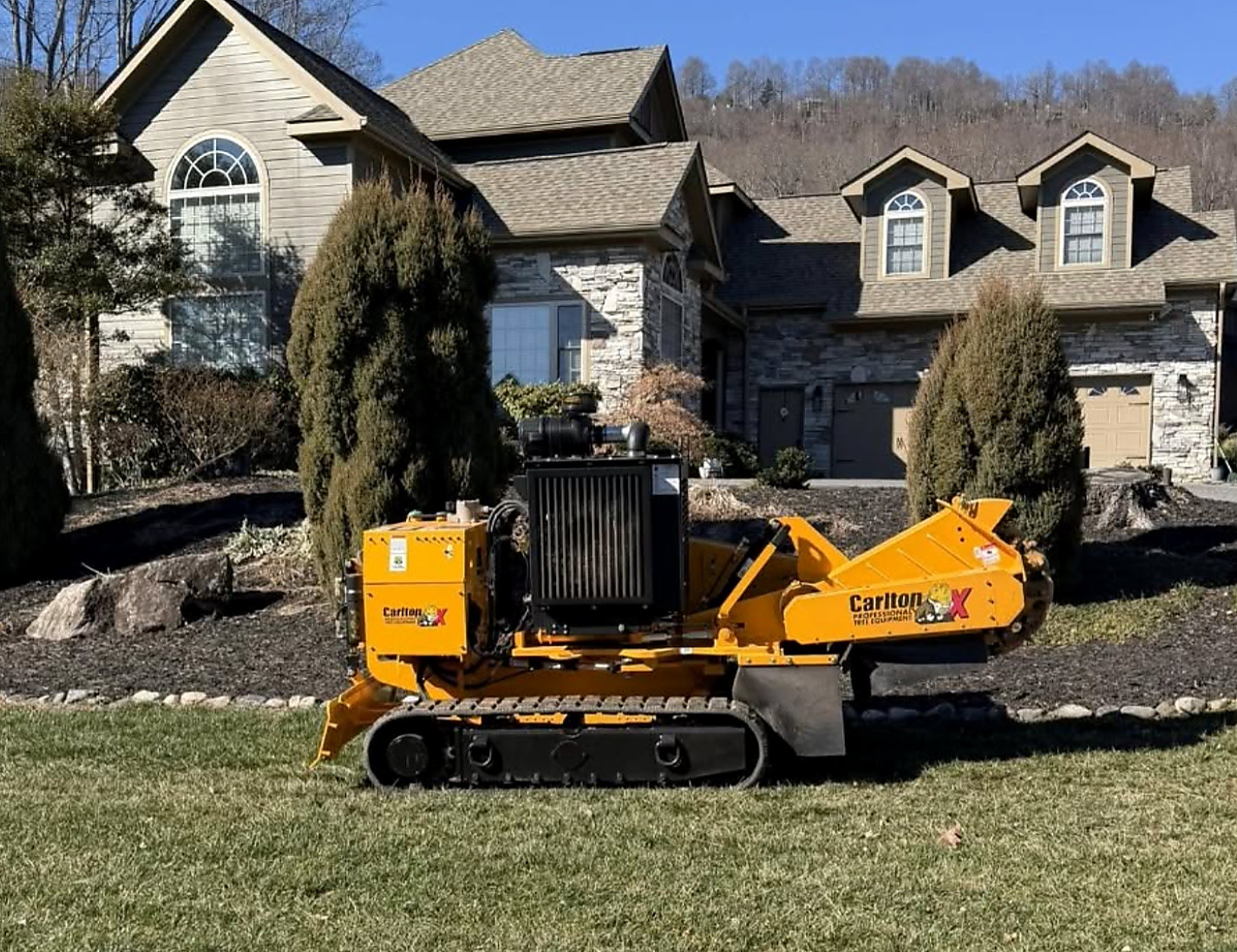 Tracked grinder on a front lawn in front of a stone-fronted home